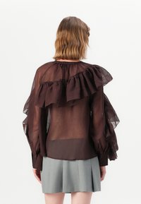 Bruine doorschijnende blouse met opvallende ruches aan de mouwen en de halslijn. Voorzien van knoopmanchetten en een relaxte pasvorm. Grijze geplooide rok eronder.