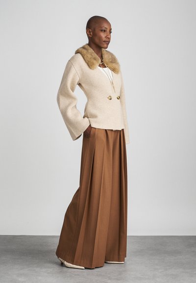 Veste beige en tricot avec col en fourrure, boutons dorés à double boutonnage, associée à un pantalon large plissé marron, sur fond gris.