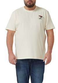 Mann trägt ein cremefarbenes T-Shirt mit einer kleinen Grafik eines laufenden Vogels im roten Pullover auf der linken Brust, kombiniert mit blauen Jeans.