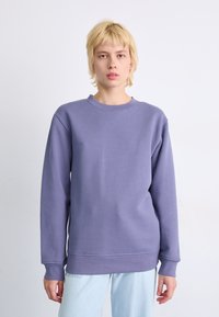 Paarse sweatshirt met een ronde hals, lange mouwen en geribbelde boorden. De stof heeft een zachte textuur. Draagt met lichtblauwe jeans.
