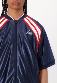 Námořnická bunda na zip s červenými a bílými akcenty, která má prodyšný síťovaný design a logo Adidas na levé části hrudi.