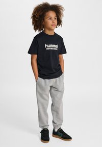 Svart t-shirt med korta ärmar och vitt "hummel"-logotyp, kombinerad med ljusgråa sweatpants och svarta sneakers med gröna detaljer.