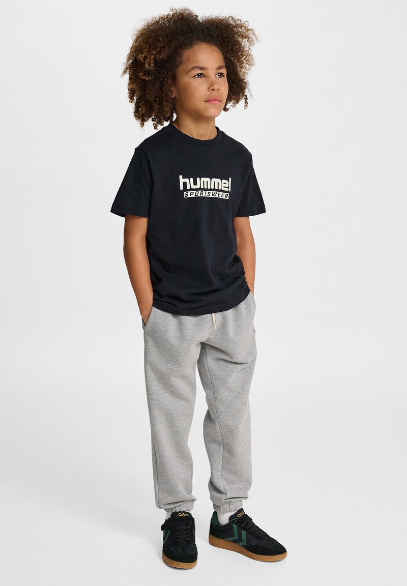 Svart t-shirt med korta ärmar och vitt "hummel"-logotyp, kombinerad med ljusgråa sweatpants och svarta sneakers med gröna detaljer.