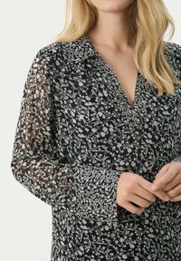 Mornarica bluza s cvjetnim uzorkom, s V-izrezom i prozirnim dugim rukavima. Istaknuti detalji uključuju kontrastni cvjetni uzorak na manžetama.