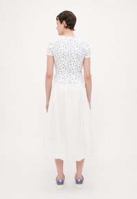 Witte top met korte mouwen en blauw bloemenpatroon, gecombineerd met een lange witte rok en paarse sneakers, gezien vanaf de achterkant.
