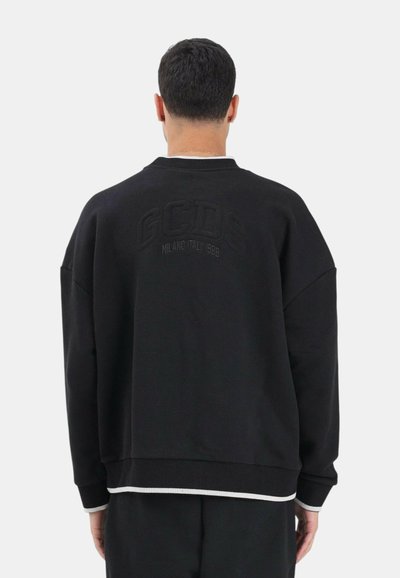 Uomo con felpa nera con la scritta "GCDS Milano Italia 1988" ricamata sulla schiena, di spalle su uno sfondo neutro.