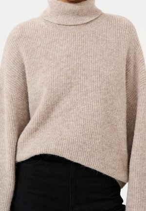 Personne portant un pull à col roulé côtelé beige partiellement rentré dans un pantalon noir taille haute sur fond blanc.