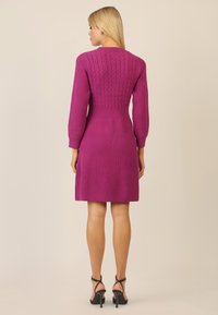Fuchsia Strickkleid mit einem Zopfmuster am Oberteil, langen Ärmeln und einem ausgestellten Rock. Kombiniert mit schwarzen Riemchenheels.