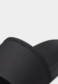 Μαύρο σανδάλι με φαρδιά λουρί, διακοσμημένο με ανάγλυφο το λογότυπο Calvin Klein επαναλαμβανόμενα.