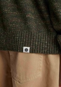 Maglione verde lavorato a maglia con orlo a coste, caratterizzato da una piccola etichetta bianca con un logo nero. Abbinato a pantaloni beige che mostrano un dettaglio della tasca.