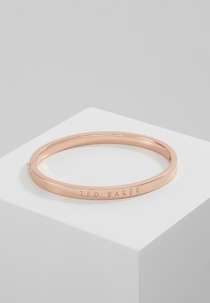 Bransoletka z metalu w różowozłotym odcieniu z wygrawerowanym napisem „TED BAKER”, umieszczona na białym geometrycznym piedestale na jednolitym szarym tle.