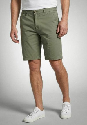Man draagt olijfgroene knielange shorts met knoop- en ritssluiting en witte lage sneakers op een grijze achtergrond.