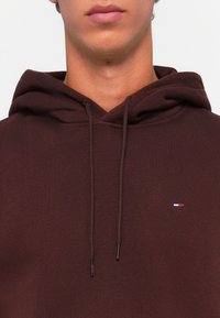Maroon hoodie with a front pocket, drawstring hood, and a small logo featuring red, white, and blue accents on the chest. Soft texture.