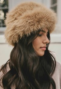 Faux fur hat in light brown, rounded design with a fluffy texture. Worn by a person with long dark hair, showcasing its soft, voluminous appearance.