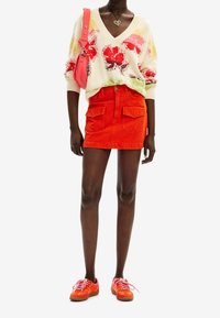 Pull en coton oversize avec motif floral en crème, orange et vert ; mini-jupe en jean rouge avec poches avant ; baskets orange avec accents roses.