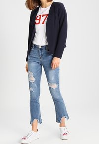 Person som bär en marinblå kavaj, vit t-shirt med "97" i röd färg, trasiga blå cropped jeans och vita sneakers med blommigt broderi.