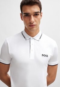 Polo blanco con ribetes negros y cierre de botones en la parte delantera, que presenta un logo bordado en el lado izquierdo del pecho que dice "BOSS". Tejido suave y ligero.