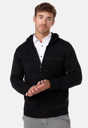 Schwarzer gestrickter Kapuzenpullover mit Reißverschluss, ausgestattet mit gerippten Bündchen und Saum, dezentem Strukturmuster und einem Etikett an der unteren Seite.
