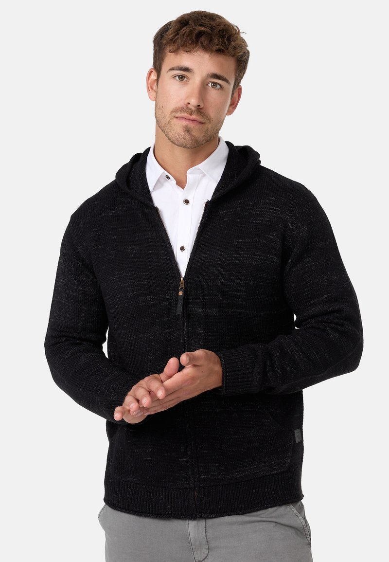 Schwarzer gestrickter Kapuzenpullover mit Reißverschluss, ausgestattet mit gerippten Bündchen und Saum, dezentem Strukturmuster und einem Etikett an der unteren Seite.