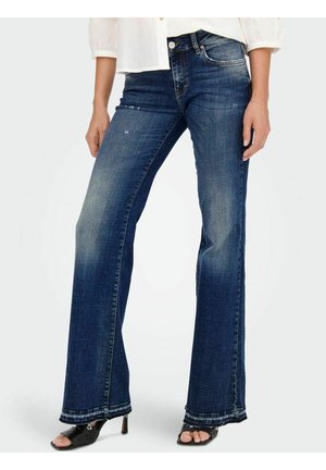 Vrouw die donkerblauwe flared jeans draagt met lichte slijtage, gecombineerd met zwarte open-toe hakken en een witte blouse met pofmouwen.