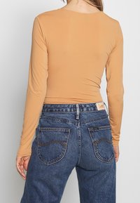 Langärmliges Oberteil aus beigem Stoff, kombiniert mit blauen Jeans aus Denim, die große Gesäßtaschen und kontrastierende Nähte aufweisen. Lee-Branding am Bund.