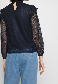 Námořnická modrá blůza s krajkou, která má průhledné dlouhé rukávy a detail v oblasti zad ve tvaru klíčové dírky. Tkanina má jemný texturovaný vzor.