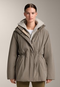 Vrouw met naar achteren gekamde haar draagt een beige geruit jasje over een licht ritsvest en olijfgroene broek tegen een effen achtergrond.