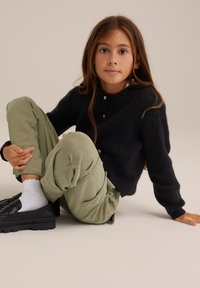 Melna, pūkaina dzija džemperis ar apaļu kaklu un trim pogām, apvienots ar olīvu zaļajiem kravas biksēm. Melni loafers un baltas zeķes noslēdz apģērbu.