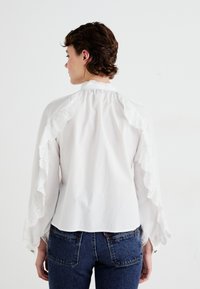 Blusa blanca con un borde de encaje bordado y volantes en los hombros y puños; ajuste entallado combinada con jeans de denim azul.
