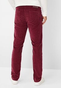 Bordeaux kleurige corduroy broek met verticale ribbels, twee achterzakken en een label op de tailleband. Gecombineerd met witte sneakers, gezien van achteren.