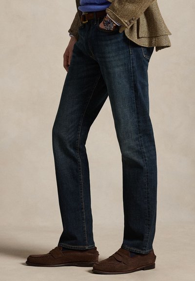 Muž nosiaci tmavomodré džínsy, hnedé semišové mokasíny, modrý sveter, hnedý vzorovaný blejzer a náramkové hodinky, ruka v vrecku.