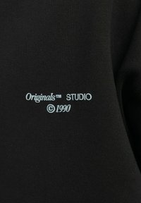 Czarna tkanina z jasnoniebieskim haftowanym napisem: "Originals™ STUDIO ©1990." Gładka struktura z minimalnymi elementami projektu.