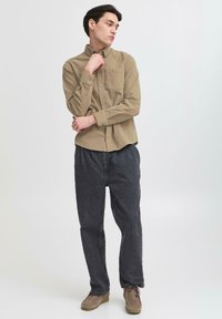 Camicetta marrone chiaro con colletto e taschino frontale, abbinata a pantaloni larghi grigi scuri e scarpe marrone chiaro su uno sfondo neutro.