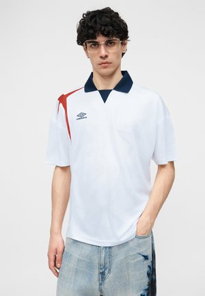 Giovane uomo con capelli ricci indossa occhiali trasparenti, maglietta sportiva bianca Umbro con colletto blu navy e accento rosso, e jeans azzurri slavati.