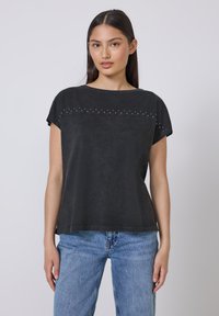 Sort kortærmet top med rund halsudskæring, tekstureret stof og dekorative nitter langs den øverste front. Sammen med lyseblå jeans.