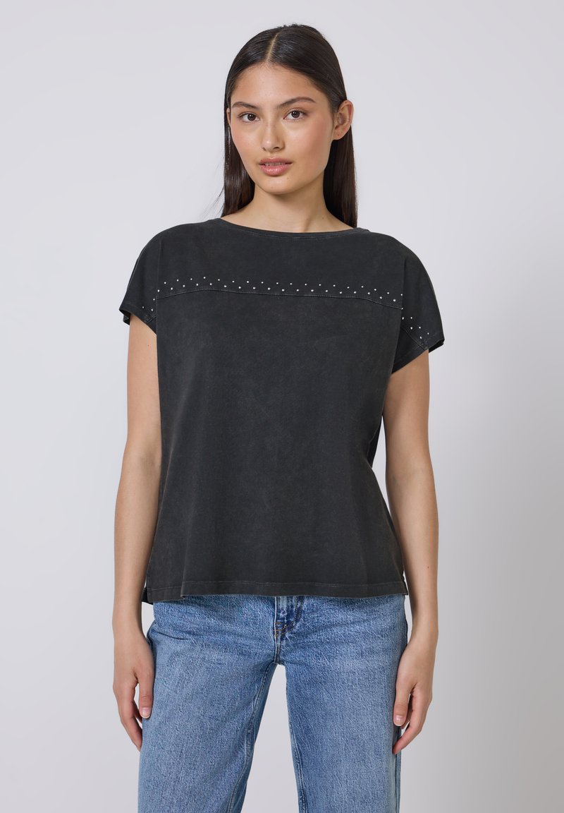 Sort kortærmet top med rund halsudskæring, tekstureret stof og dekorative nitter langs den øverste front. Sammen med lyseblå jeans.