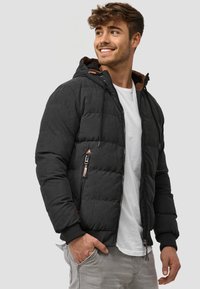 Schwarze Pufferjacke mit matter Oberfläche, Reißverschluss, verstellbarer Kapuze und elastischen Bündchen. Mit seitlichen Reißverschlusstaschen und braunen Akzenten.