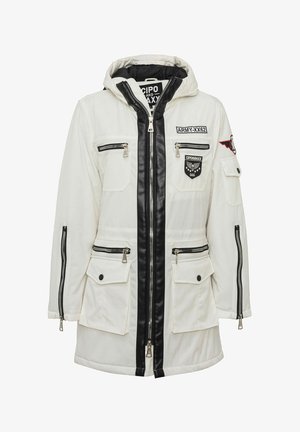 Witte parka met capuchon, zwarte ritstrek, meerdere zakken met rits en knopen, en geborduurde patches op borst en mouw.