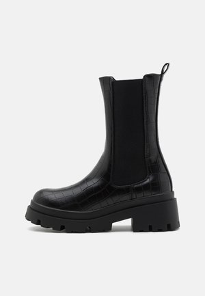 Botte noire mi-mollet avec semelle épaisse, panneau latéral élastique, languette à l’arrière et texture crocodile sur la tige en cuir.
