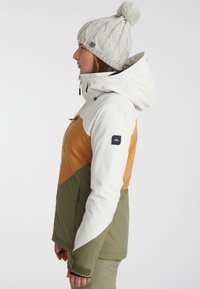 Women's ski jacket in white, orange, and olive green. Features a zippered pocket, adjustable hood, and textured knit beanie with pom-pom.
