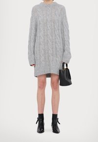 Personne portant une robe-pull ample en maille torsadée grise, des bottines noires et tenant un sac à main noir sur un fond uni.