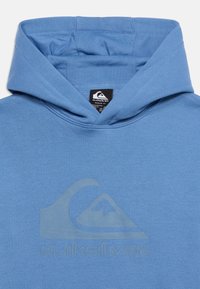 Sudadera con capucha azul de Quiksilver con logo en relieve de ola y montaña en el pecho, mostrando la capucha y la etiqueta interior de la marca.