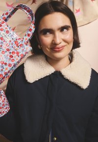 Fiatal nő rövid sötét hajjal, sötétkék kabátot visel krémszínű gyapjúgallérral, kezében virágmintás táskával, amely piros, lila és narancssárga virágokat ábrázol.