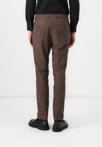 Pantaloni maro texturat cu un model subtil, buzunare pe partea din spate și tivuri răsucite, asortați cu pantofi negri.