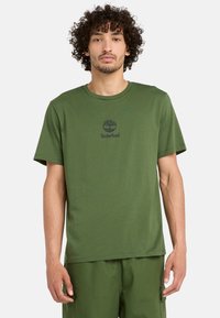 Grön bomullströja med korta ärmar, rund halsringning, med en mörk logotyp ovanför bröstet. Mjuk textur, avslappnad passform, enkel design.