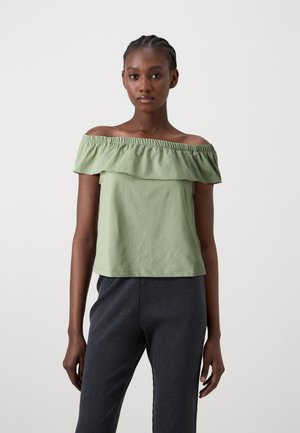 Grön off-shoulder topp med ett volangkrage, tillverkad av mjukt tyg, kombinerad med mörka ruggade byxor.
