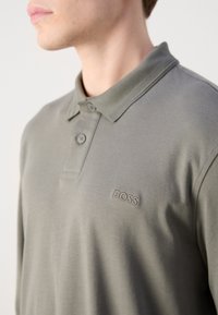 Polo majica od maslinasto zelene tkanine s ovratnikom, plaketom na dva gumba i izvezenim "BOSS" logotipom na lijevoj strani prsnog koša. Teksturedni ovratnik.