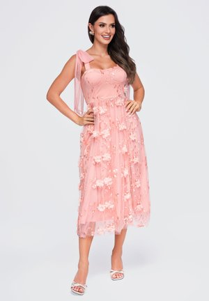 Femme souriante, portant une robe midi fleurie rose sans manches avec des bretelles à nœud et des sandales blanches à talons, mains sur les hanches.