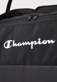 Black fabric bag features a smooth texture with a prominent white "Champion" logo. Dual zipper pulls enhance functionality.