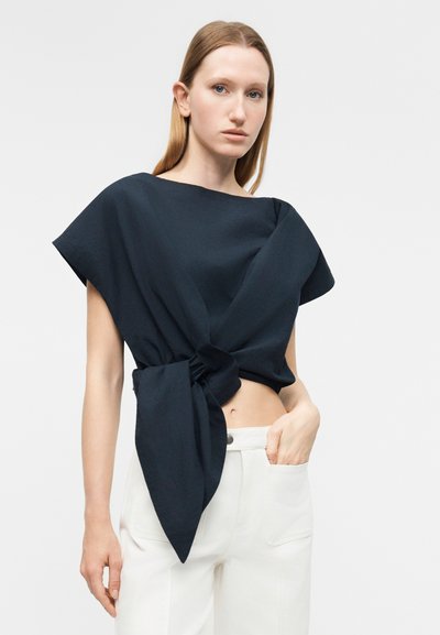 Γυναίκα που φορεί ένα ασύμμετρο μπλε ναυτικό τοπ δεμένο στη μέση και λευκό ψηλόμεσο παντελόνι, πόζαρει μπροστά σε ένα απλό φόντο.